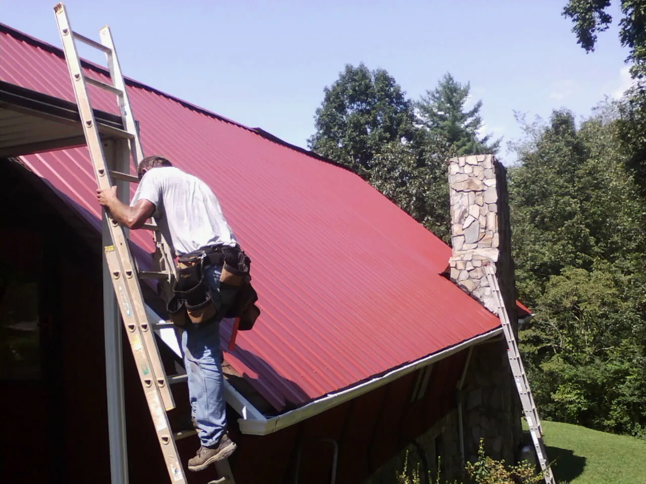 Roofer on a ladder beside a red metal roof and stone chimney — Soffit & Fascia Repair by Roofing Brothers in Waynetown
